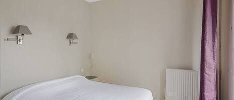 Un dormitorio con una cama, dos lámparas fijadas a la pared, un radiador blanco y una cortina morada.