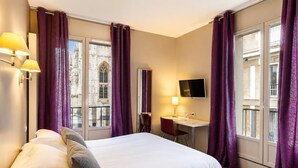 A hotel room with a bed, desk, and large windows with purple curtains.
