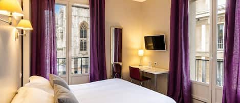 A hotel room with a bed, desk, and large windows with purple curtains.