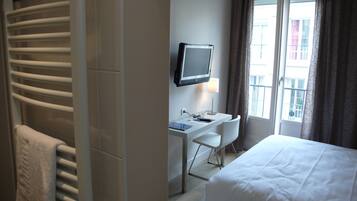 Una habitación de hotel con una cama, un escritorio con computadora y un televisor montado en la pared, una ventana con cortinas y un perchero para toallas.