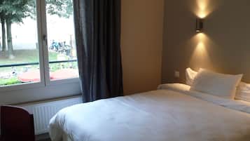 A hotel room with a large bed, a chair, a window with curtains, and a wall-mounted light.