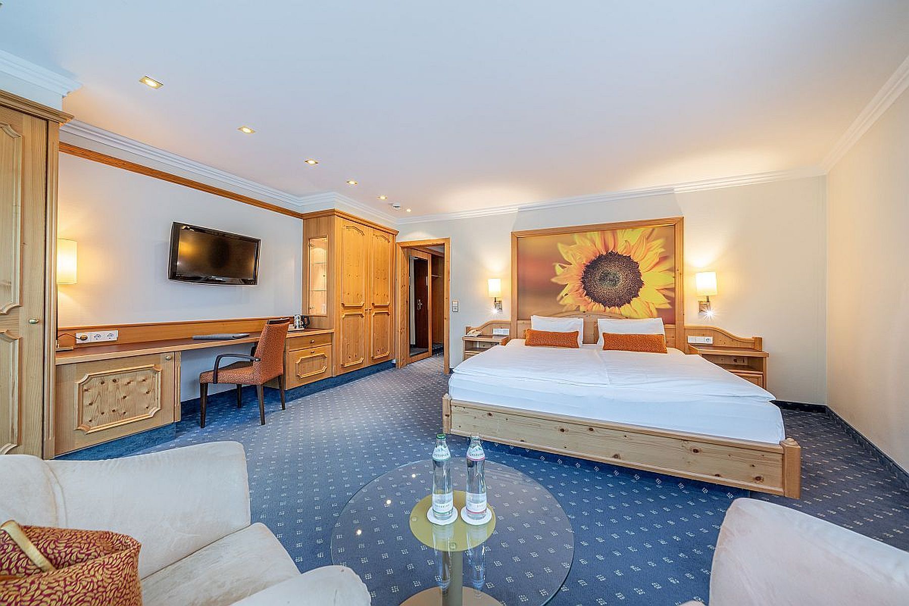 A hotel room with a bed, a desk, a chair, a TV, and a large sunflower painting.