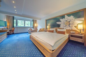 A hotel room with a large bed, a desk with a computer, a chair, and a large flower painting on the wall.
