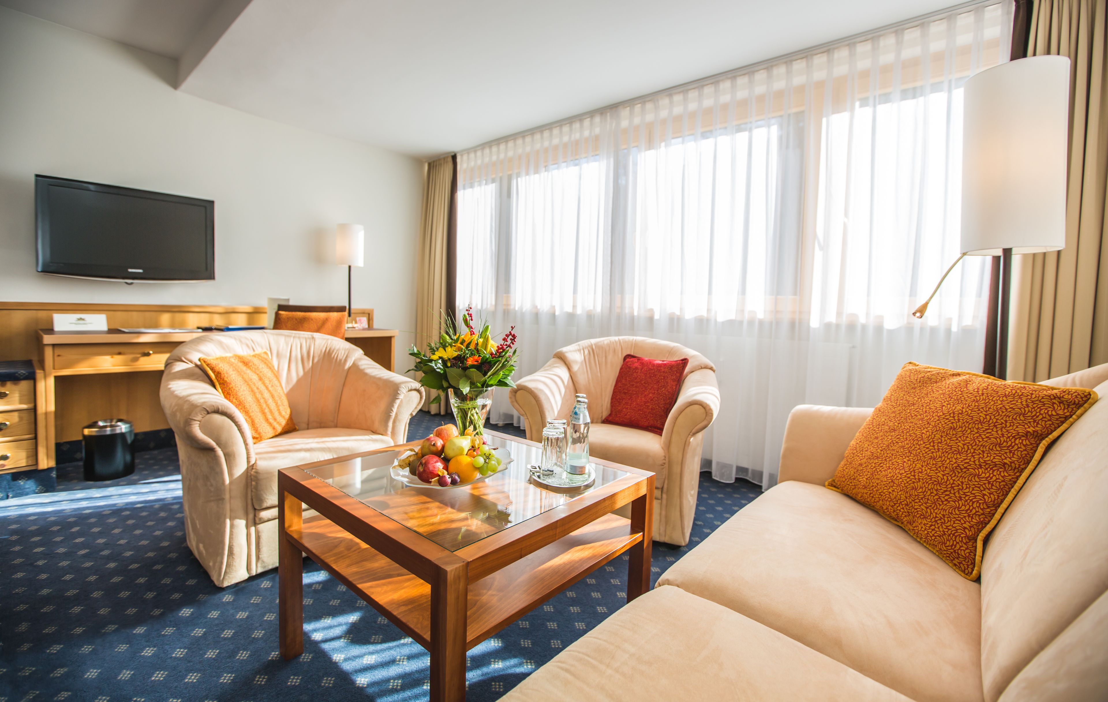 Een hotelkamer met een bank, twee fauteuils, een bijzettafeltje met fruit en een vaas, een televisie en een bureau.