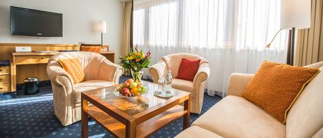 A hotel room with a sofa, two armchairs, a coffee table with fruit and a vase, a TV, and a desk.
