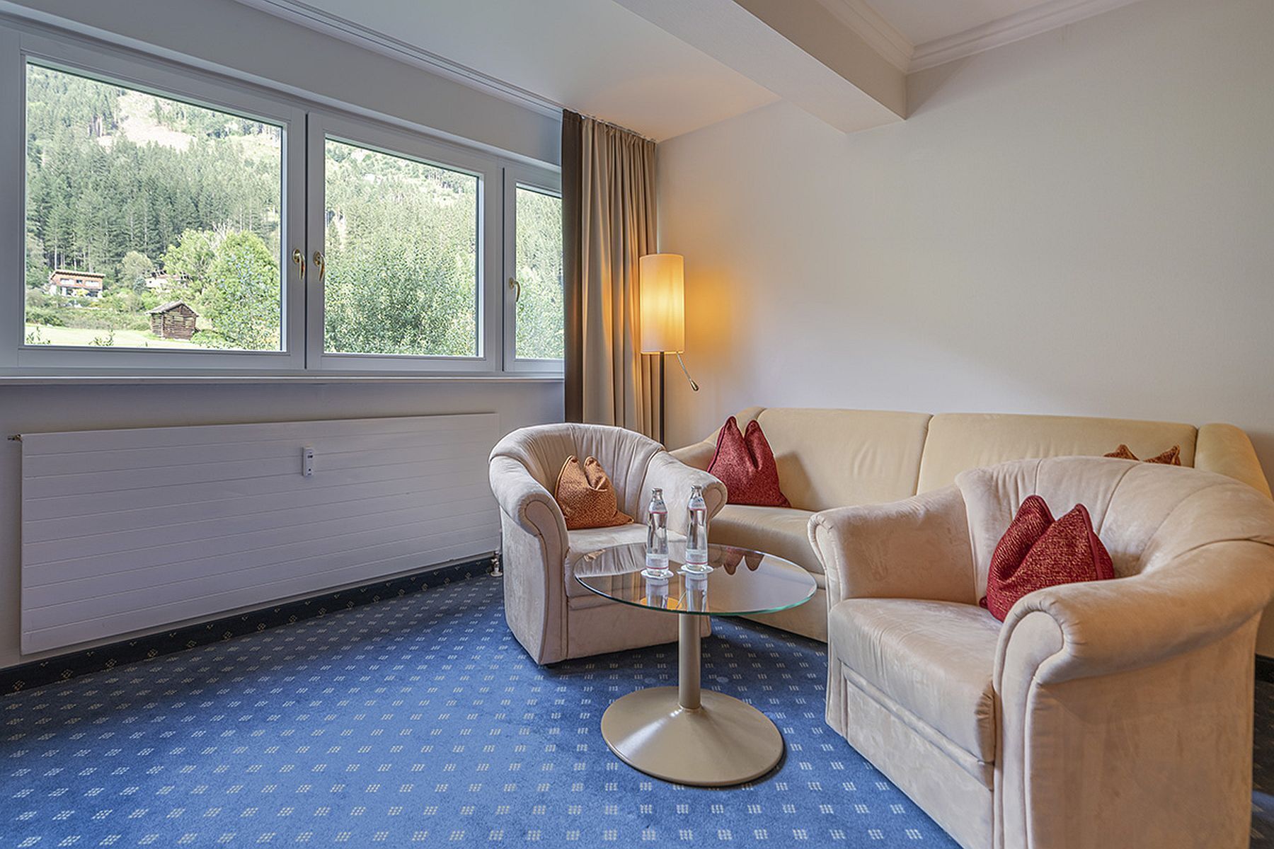 Una habitación con un sofá color beige, un sillón a juego, una mesa de centro de cristal y un ventanal con vistas a zonas verdes y un edificio.