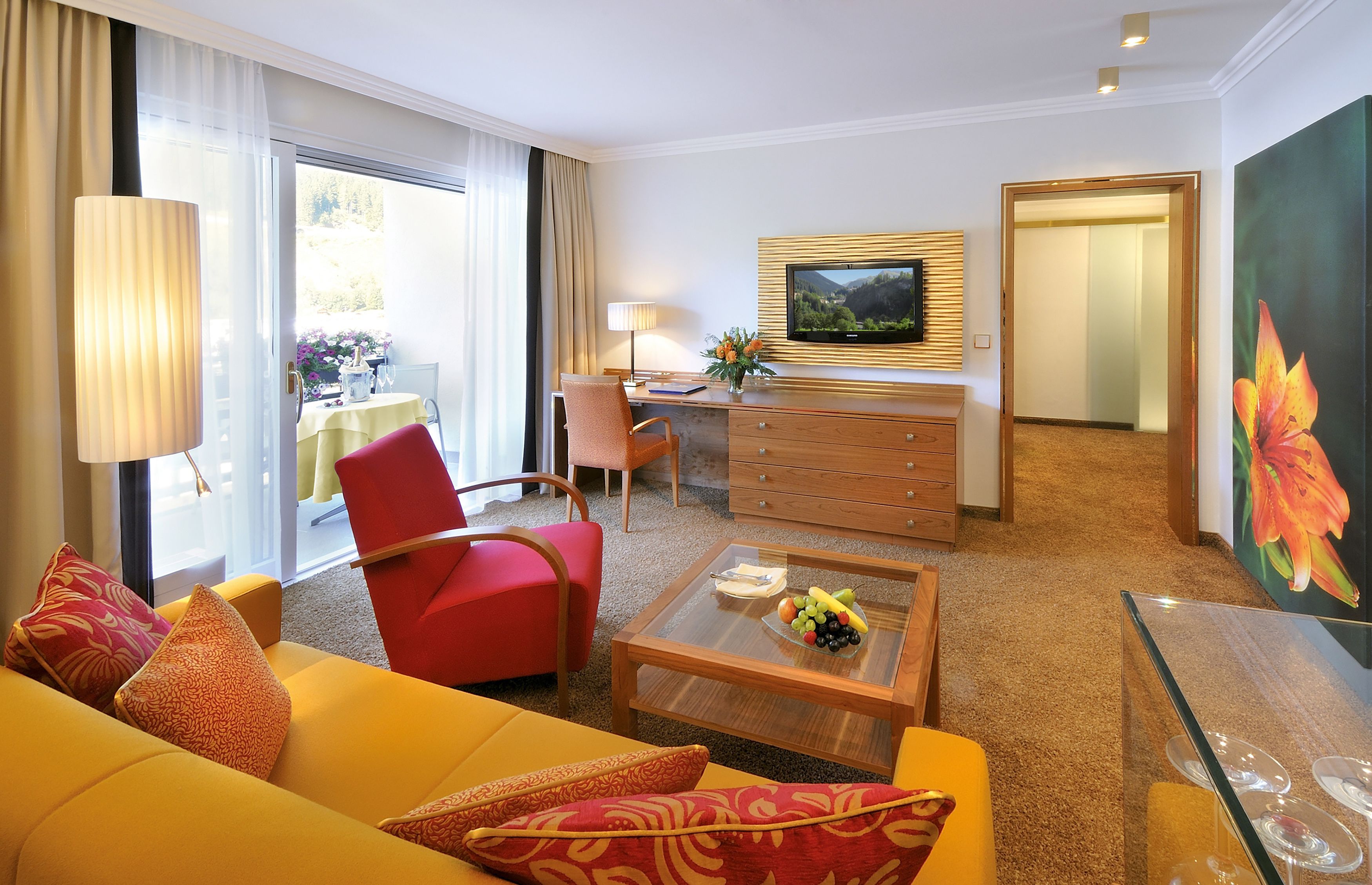 Habitación de hotel con sofá amarillo, sillón rojo y escritorio con televisor.