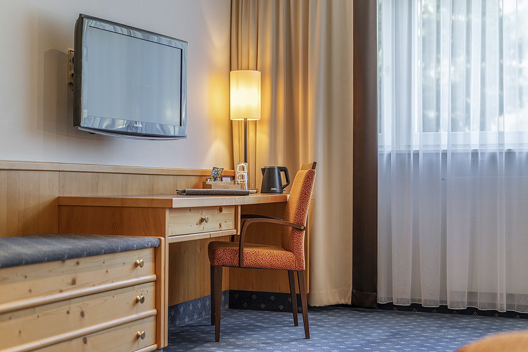 A hotel room with a wooden desk, a chair, a television mounted on the wall, a lamp, and a window with sheer curtains.