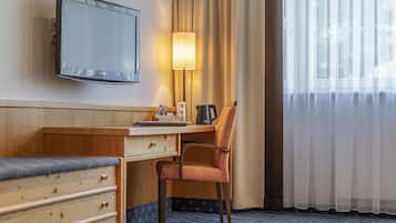 A hotel room with a wooden desk, a chair, a television mounted on the wall, a lamp, and a window with sheer curtains.