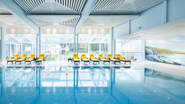 A modern indoor swimming pool with clear glass walls, yellow lounge chairs, and a large mural of a coastal landscape. - EUROPÄISCHER HOF Bad Gastein (Bad Gastein)