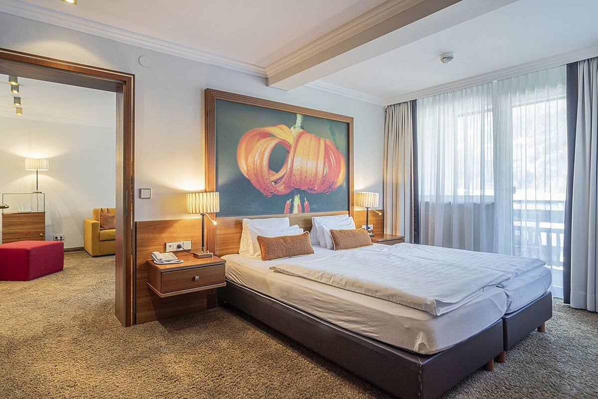 Une chambre d’hôtel avec un grand lit, deux tables de chevet, une petite table d’appoint, un pouf rouge, un grand tableau et une vue sur l’extérieur à travers des rideaux transparents.