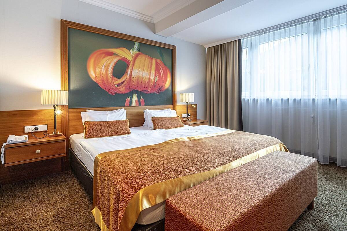 Une chambre d’hôtel avec un grand lit, deux tables de chevet, un tableau et une fenêtre avec des rideaux.