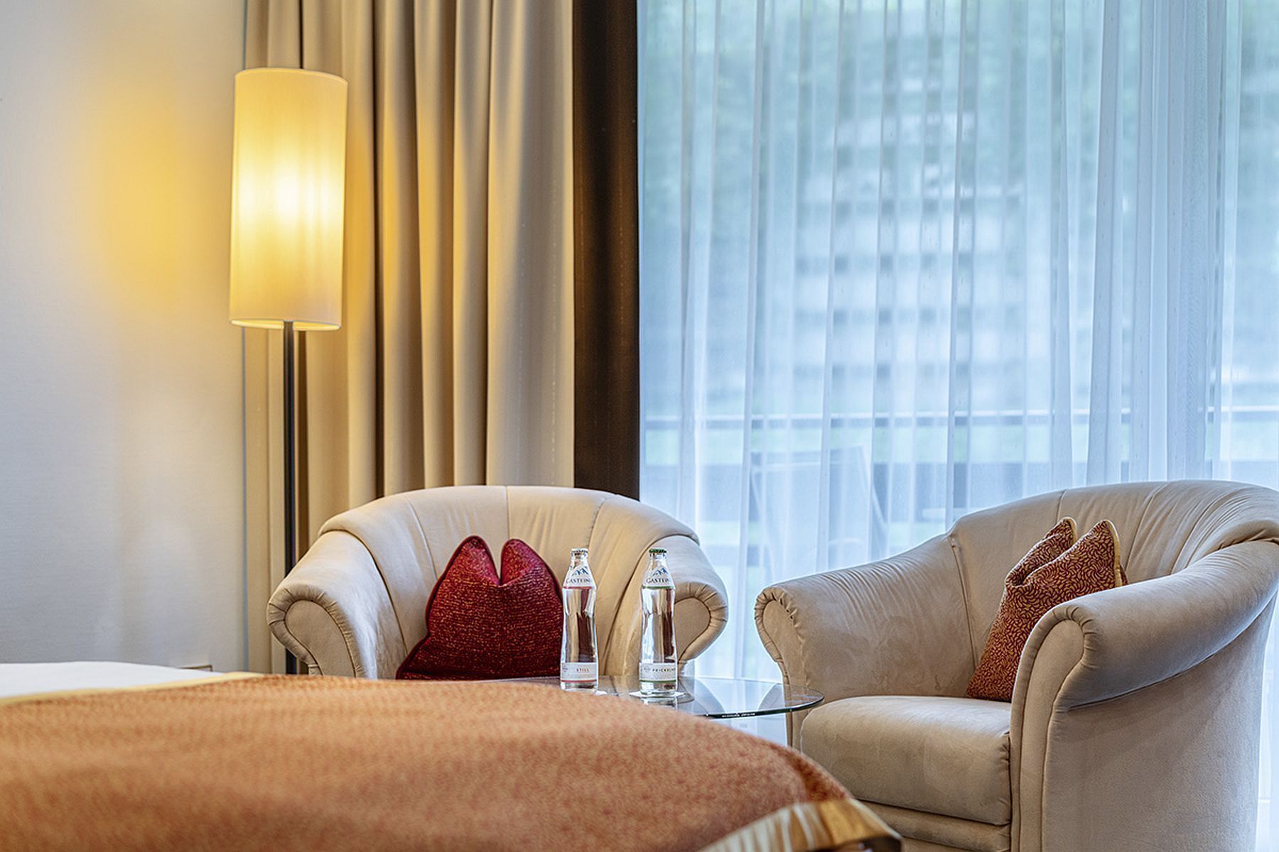 A hotel room with two beige armchairs, a glass coffee table, and a bed with a brown blanket. There is a lamp on the wall and a window with sheer curtains.