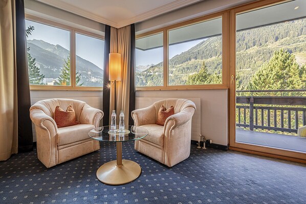 A room with two beige armchairs, a glass table, and a floor lamp. Large windows offer a view of mountains and trees.