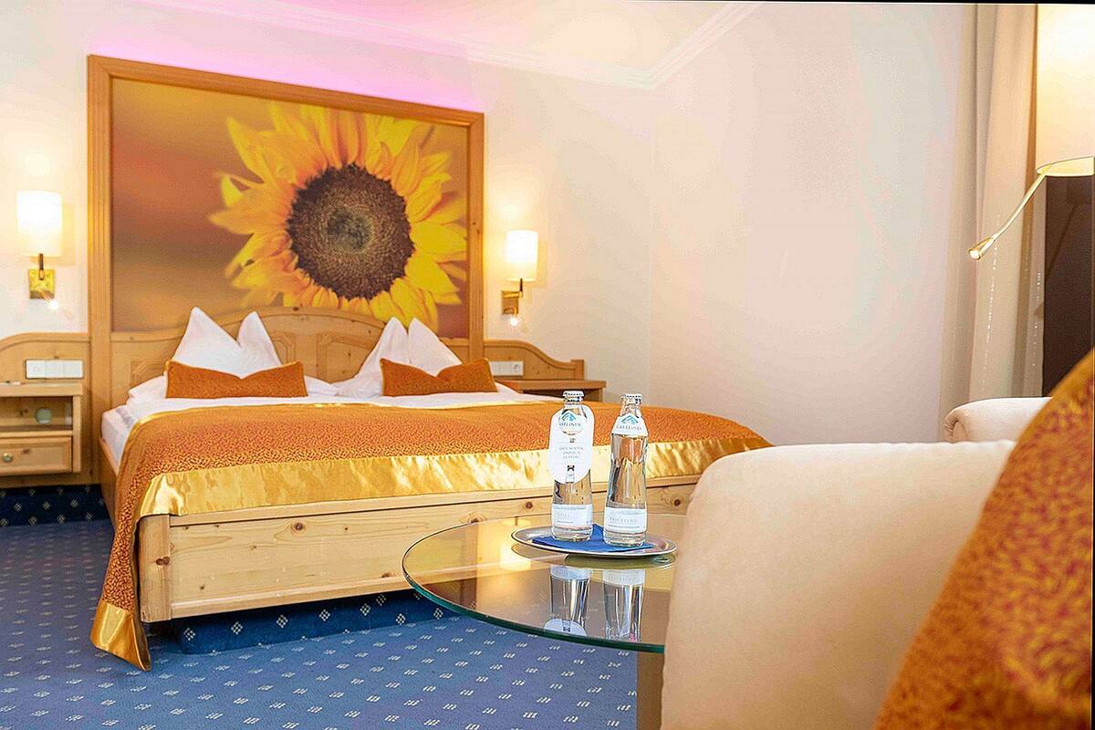 Une chambre d’hôtel avec un lit, une table en verre sur laquelle sont posées deux bouteilles, et un grand tableau représentant un tournesol accroché au mur.