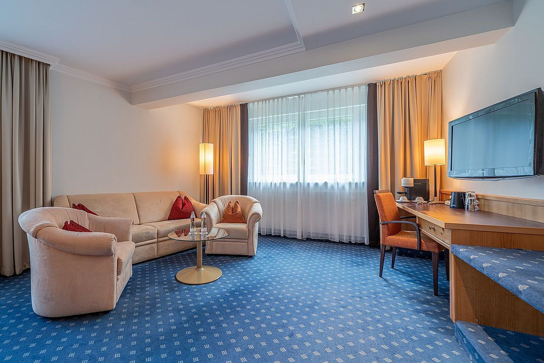 A hotel room with a sofa, armchair, and desk with a television.