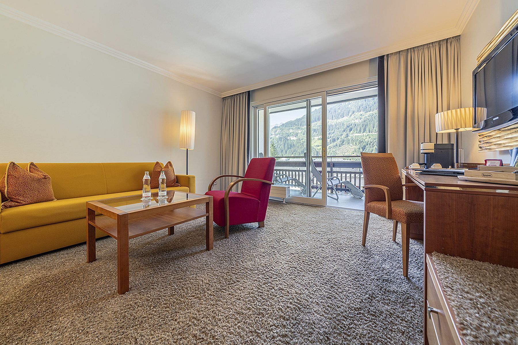 Hotelkamer met een gele bank, een rode fauteuil, een houten salontafel, een bureau met een computer en een groot raam met uitzicht op een bergachtig landschap.