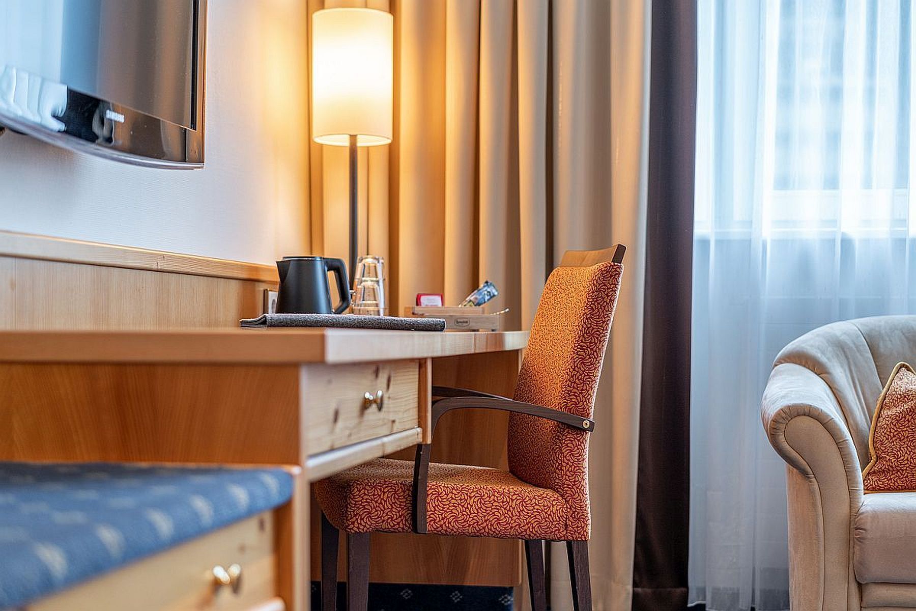 Ein Hotelzimmer mit einem Holztisch, einem gemusterten Sessel, einem weißen Sessel, einer Lampe und einem fenster mit Vorhängen.