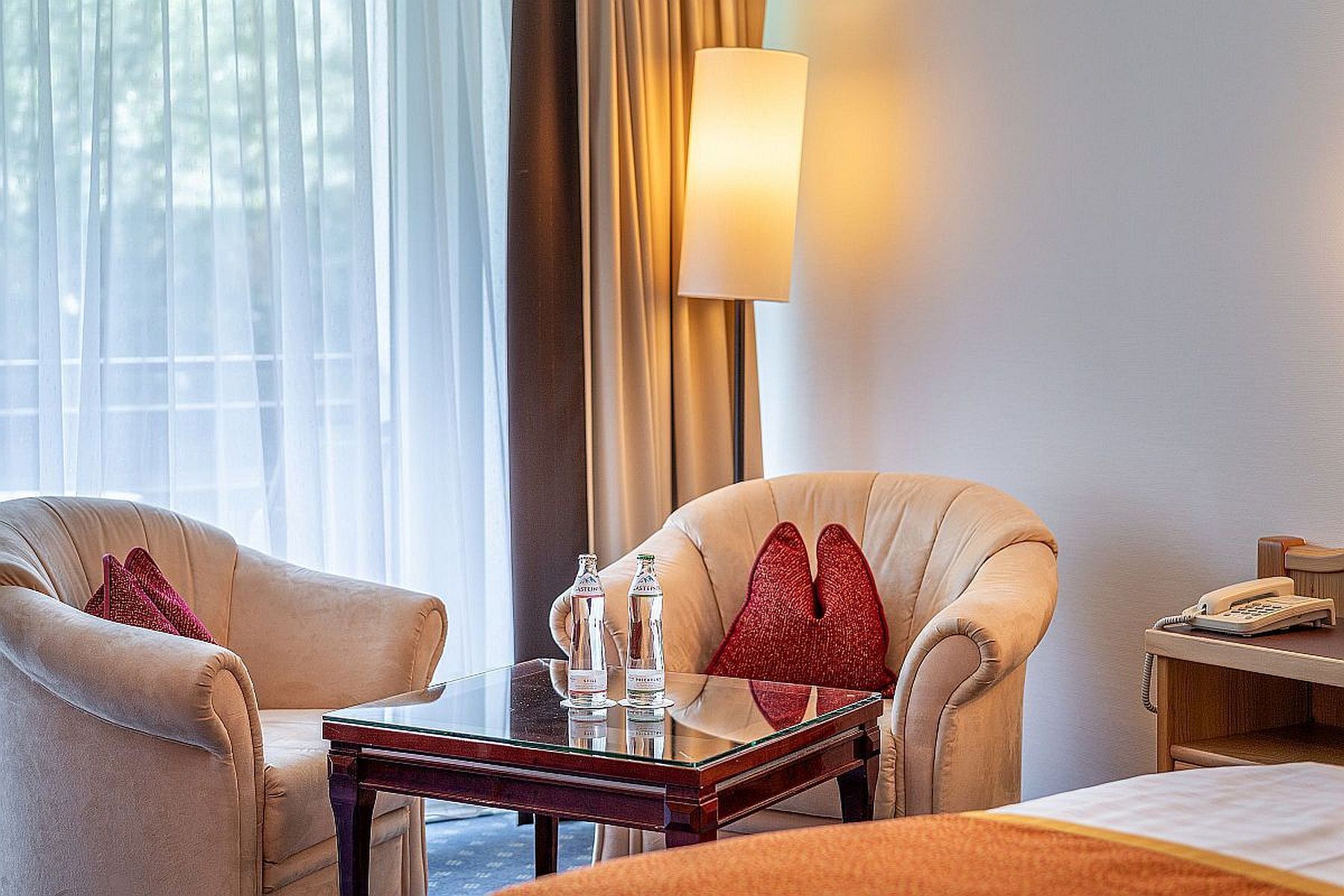 A hotel room with two armchairs, a small table with a bottle and glass, a lamp, and a bedside table with a phone.