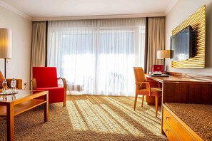 A hotel room with a large window, a desk, a red chair, a wooden dresser, and a television on the wall.