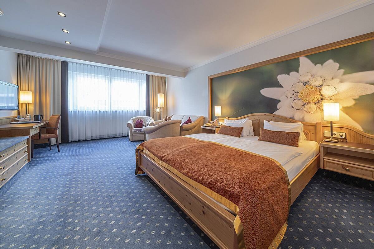 Une chambre d’hôtel avec un grand lit, un bureau, une chaise, un canapé et un grand tableau représentant des motifs floraux accroché au mur.