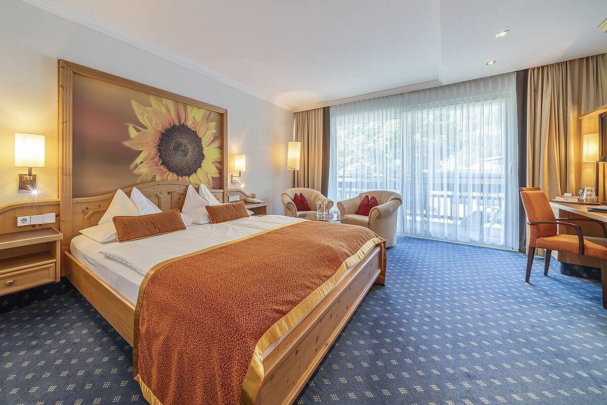Une chambre d’hôtel avec un grand lit, un bureau, une chaise, une grande fenêtre avec des rideaux et un tableau représentant un tournesol accroché au mur.