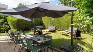 An outdoor patio with a table and chairs, a large umbrella, and a grassy area with a hedge in the background. - Le Carline, Sure Hotel Collection by Best Western (Caen)