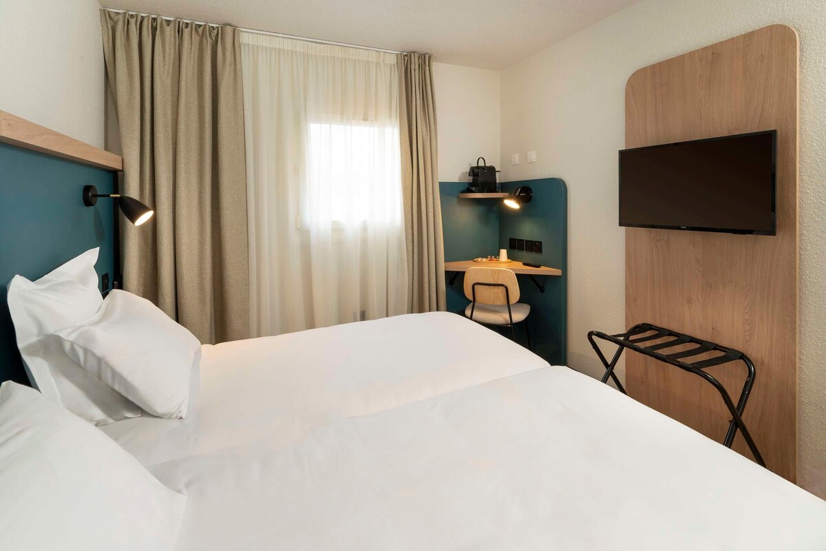 Habitación de hotel con cama, escritorio con silla, televisor montado en la pared y ventana con cortinas.