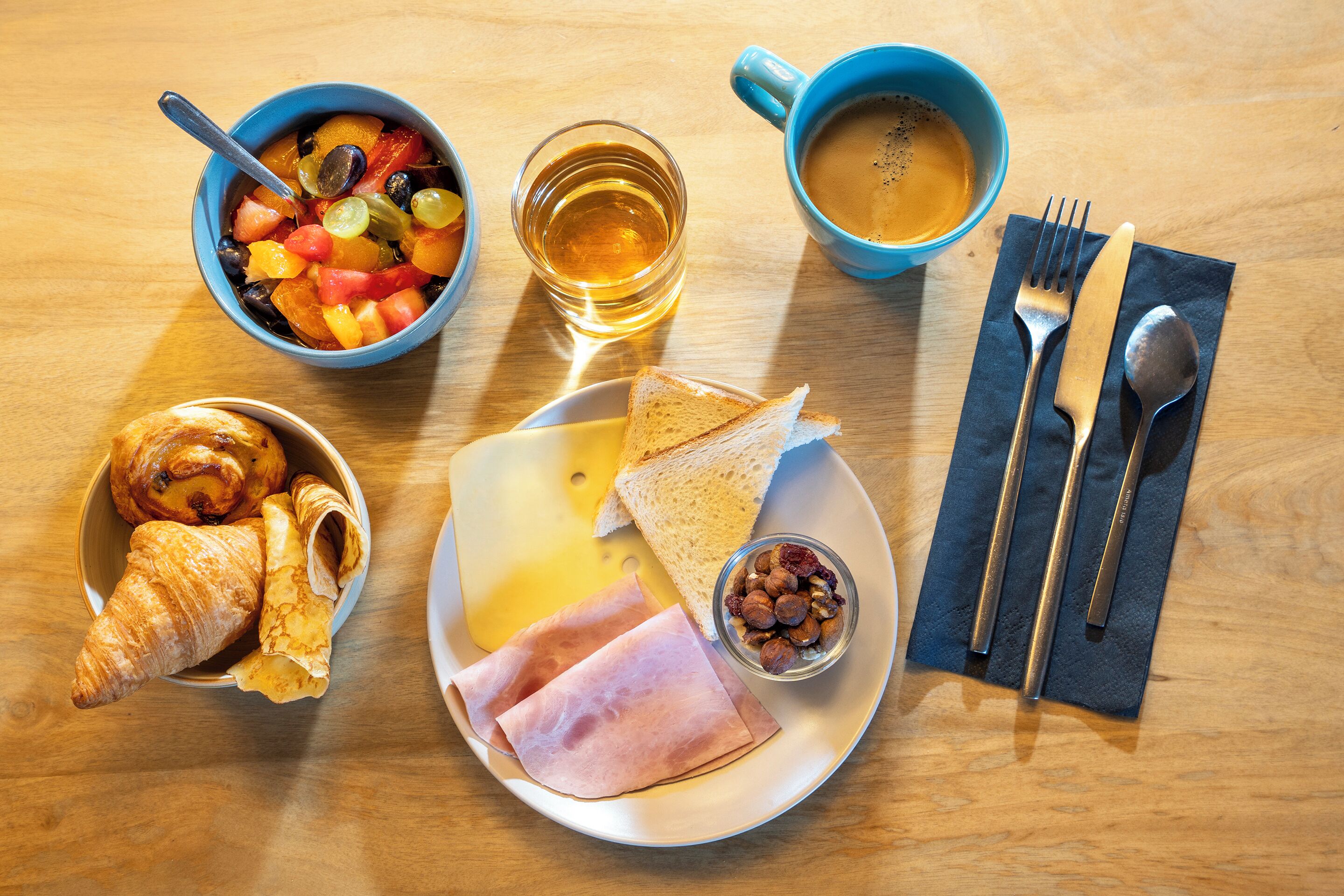 A breakfast setting with croissants, cheese, ham, toast, fruit, nuts, coffee, and tea on a wooden table.