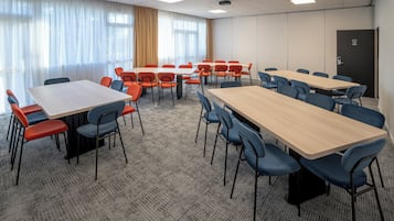 A conference room with multiple tables and chairs, large windows with curtains, and a carpeted floor.