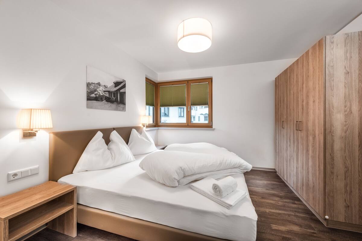 Una cama bien hecha con sábanas blancas, una mesita de noche con lámpara, un armario de madera y una ventana con persianas.