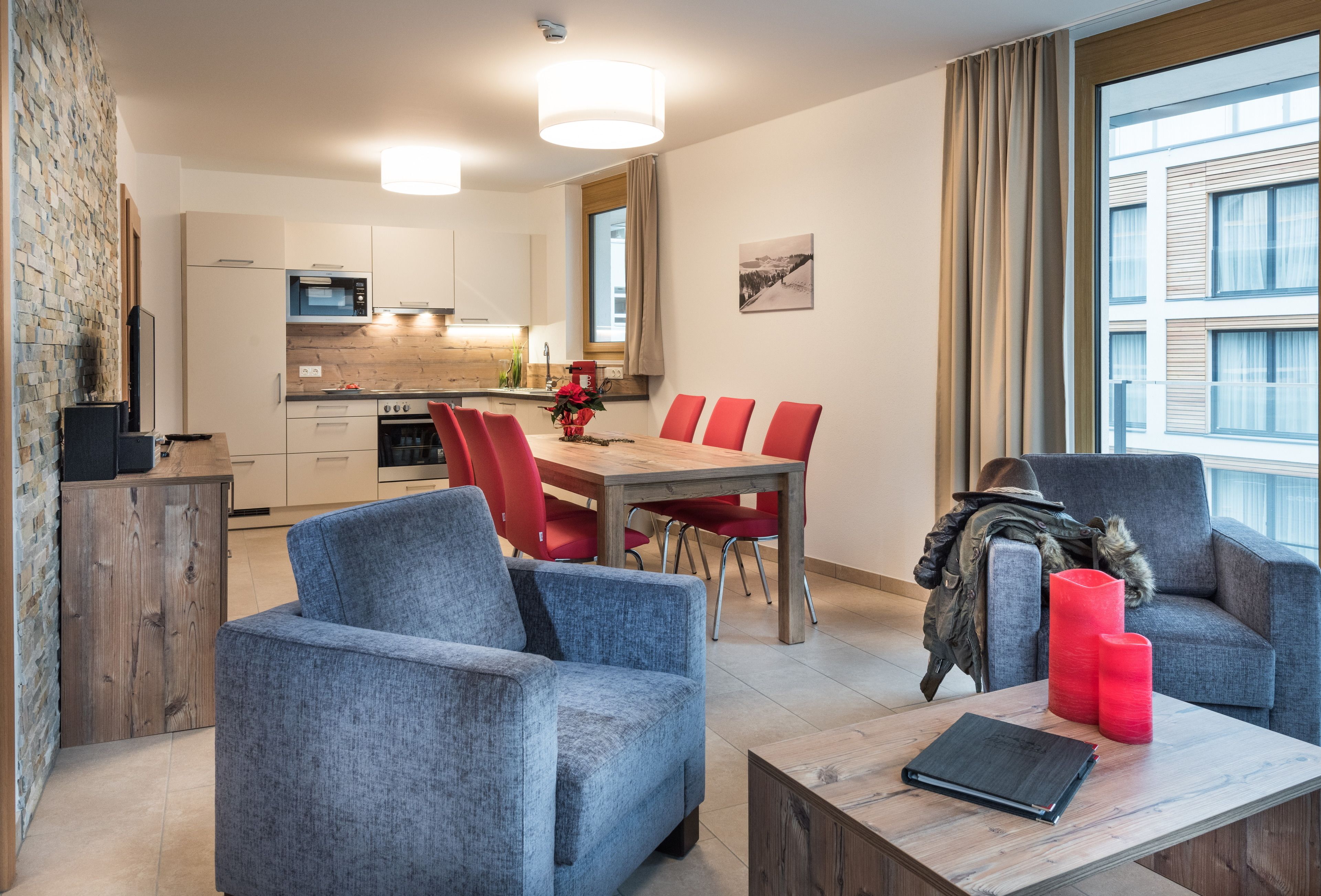 A modern living room with a grey sofa, red chairs, a wooden coffee table, and a view of the kitchen and dining area.