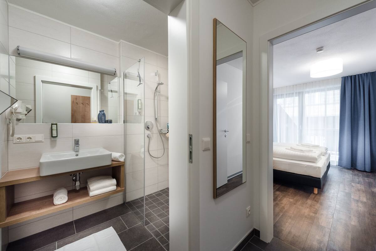 Un baño moderno con lavamanos, espejo y ducha. Junto, un dormitorio con cama y vistas al exterior.