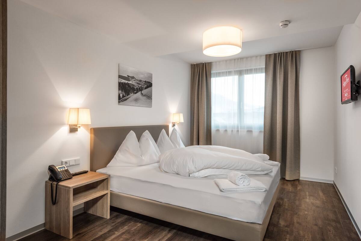 Habitación de hotel con cama, lámparas de noche, mesita de noche con teléfono, un cuadro en la pared y una ventana con cortinas.