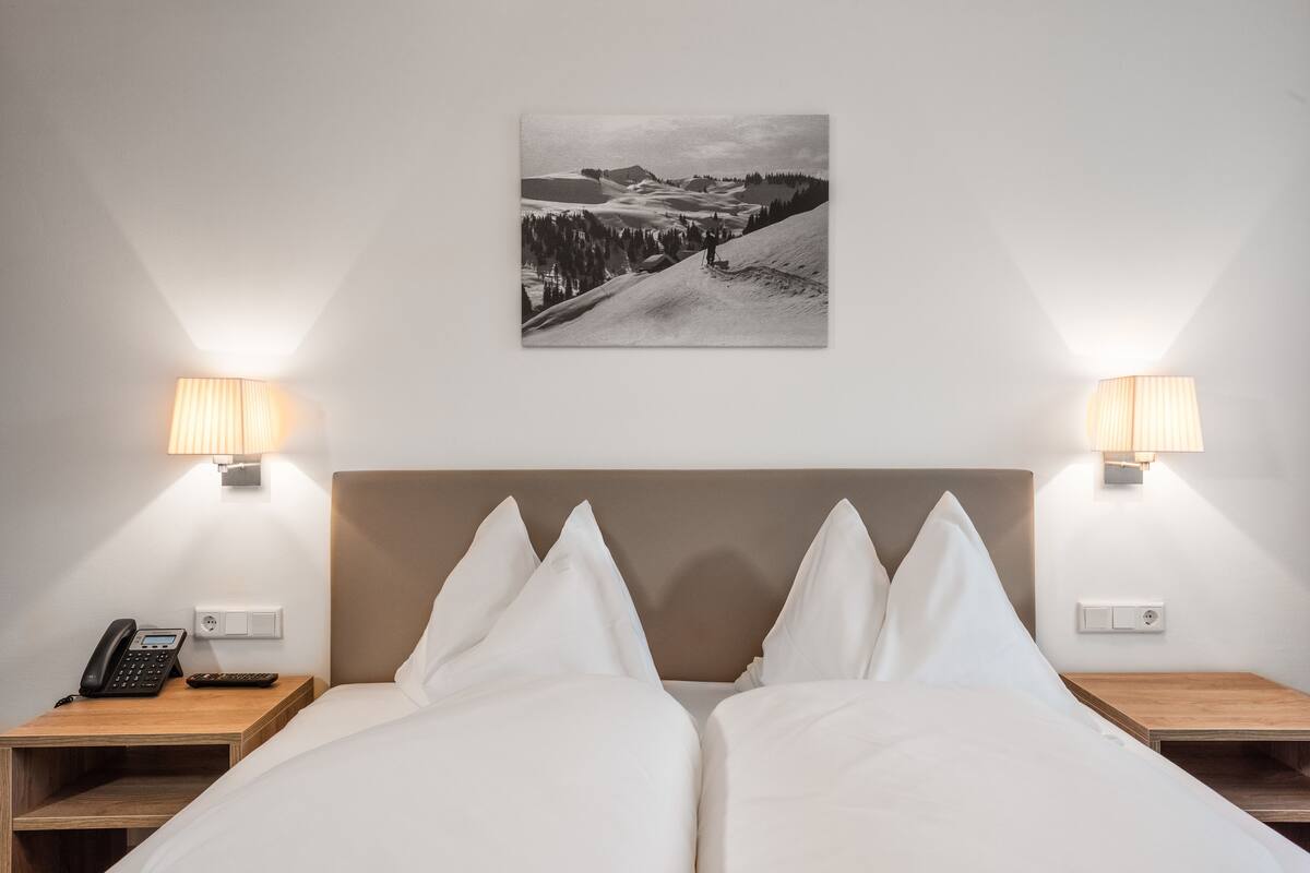 Habitación de hotel con cama, dos mesitas de noche, lámpara, teléfono y un cuadro colgado en la pared.