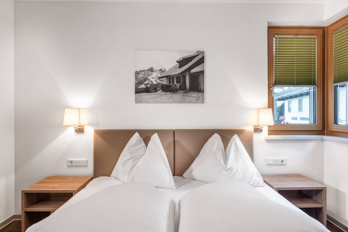 Un dormitorio con cama, dos mesitas de noche, una lámpara de pared y una ventana con persianas.