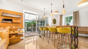 A modern kitchen with a breakfast bar, yellow chairs, and a large window. - Das Grüne Hotel zur Post (Salzburg)