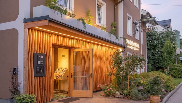 A hotel entrance with a wooden facade, a glass door, and a small garden area with flowers and plants. - Das Grüne Hotel zur Post (Salzburg)