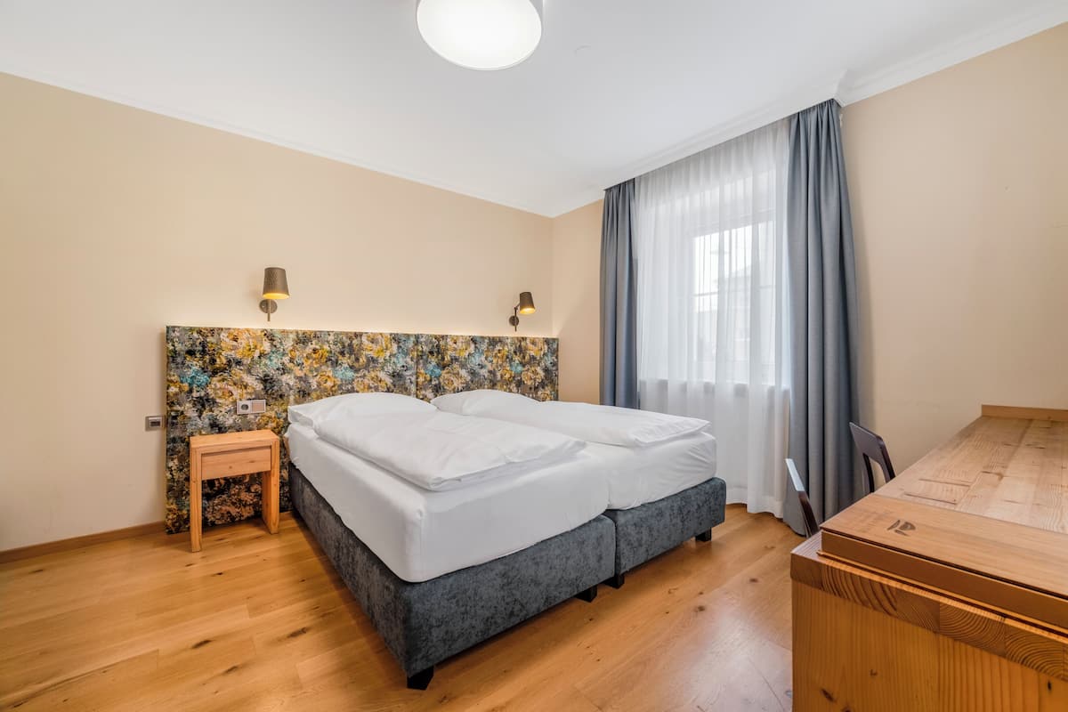 Quarto com cama, mesa de cabeceira de madeira, luminária, cabeceira com padrão floral, cômoda de madeira e janela com cortinas.