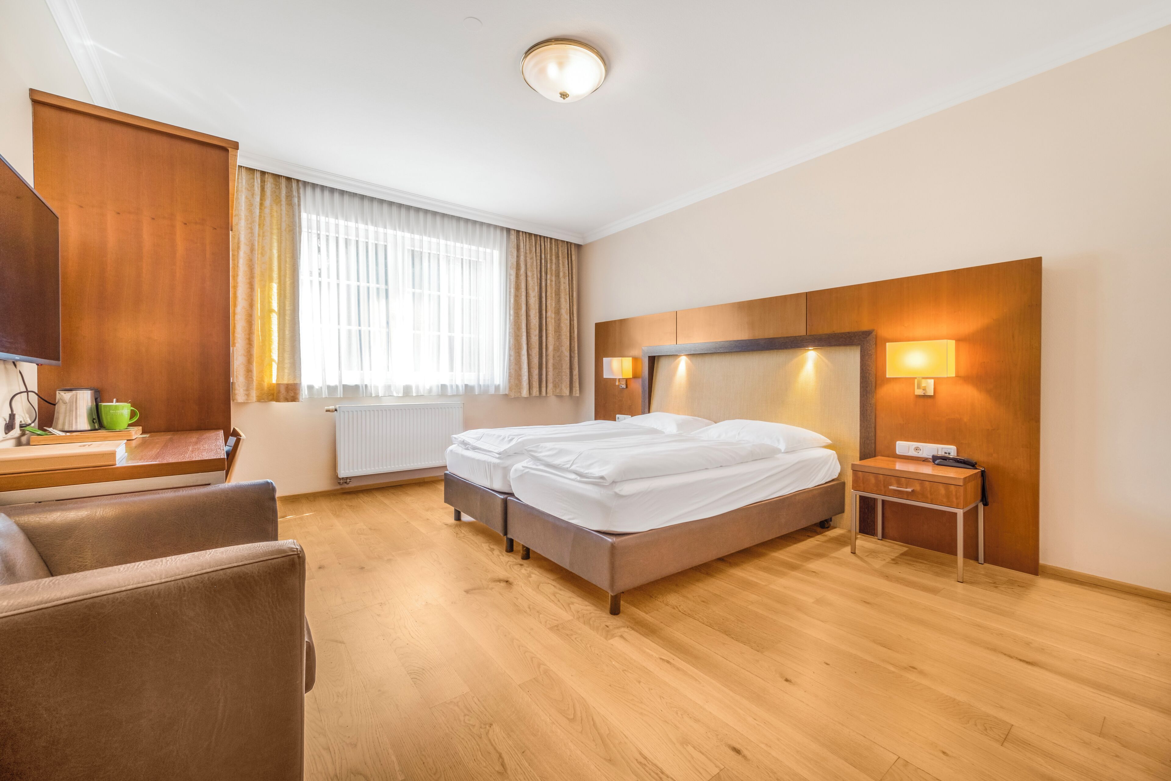 A hotel room with a bed, a desk with a lamp, a chair, and a window with curtains.