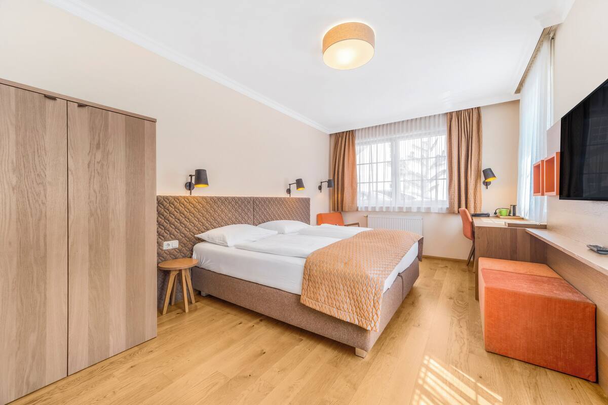 Quarto de hotel moderno com cama, escrivaninha, cadeira, guarda-roupa e janela com cortinas.