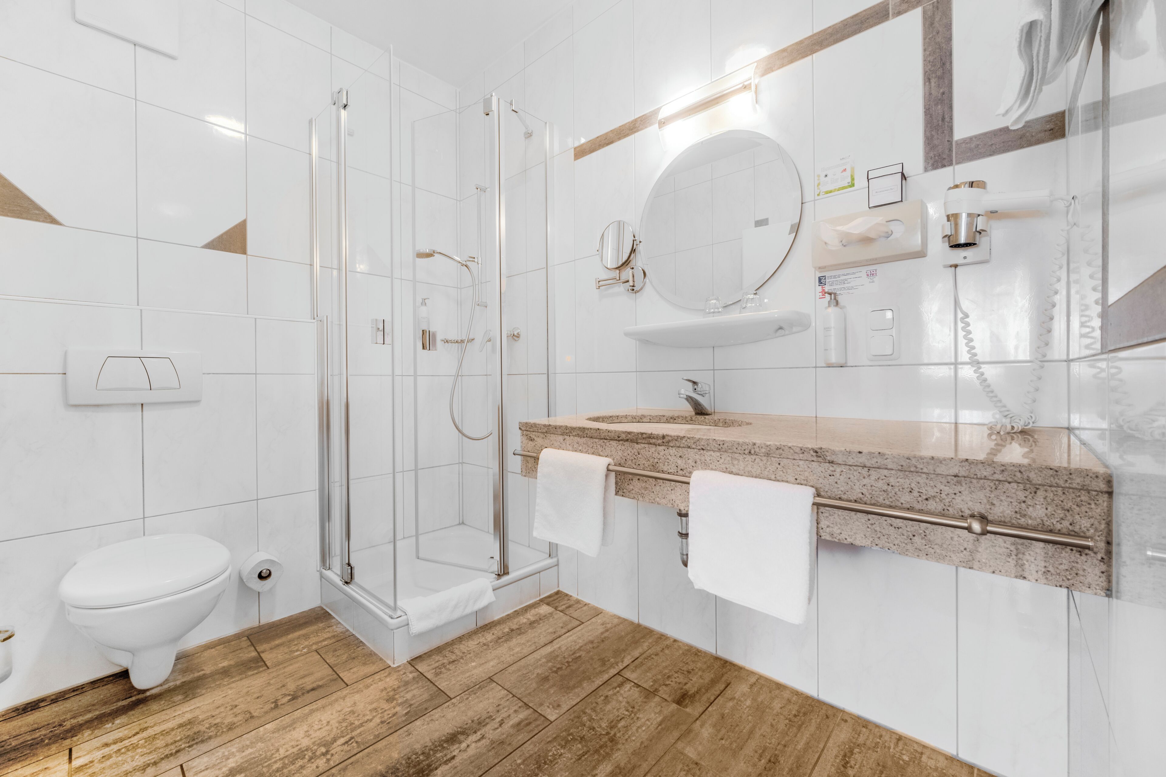 A modern bathroom with a glass shower enclosure, a white toilet, a granite countertop sink, and a mirror with a round frame.