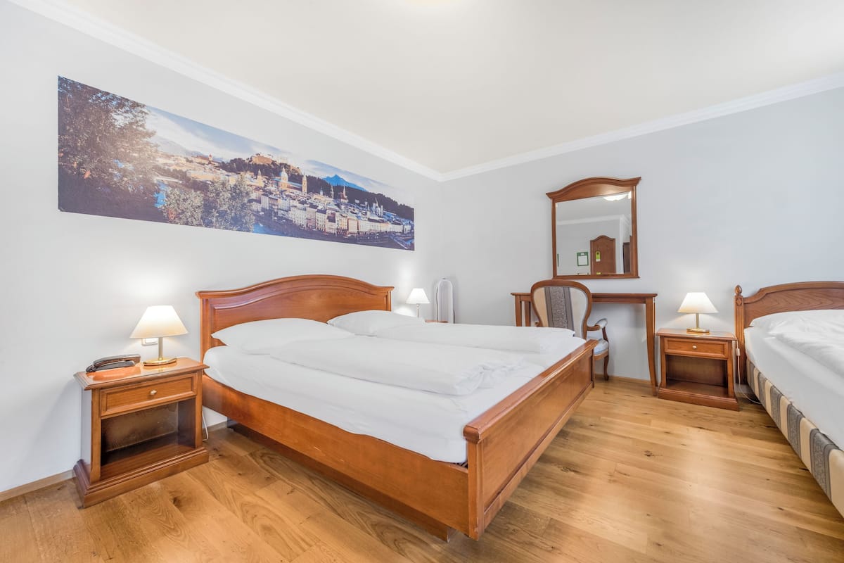 Quarto com cama de madeira, duas mesas de cabeceira com abajures, um espelho e uma pintura com vista para a cidade na parede.