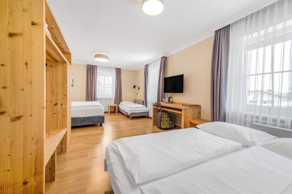 Quarto de hotel com guarda-roupa de madeira, duas camas de solteiro, uma escrivaninha com cadeira, televisão e janelas amplas com cortinas.