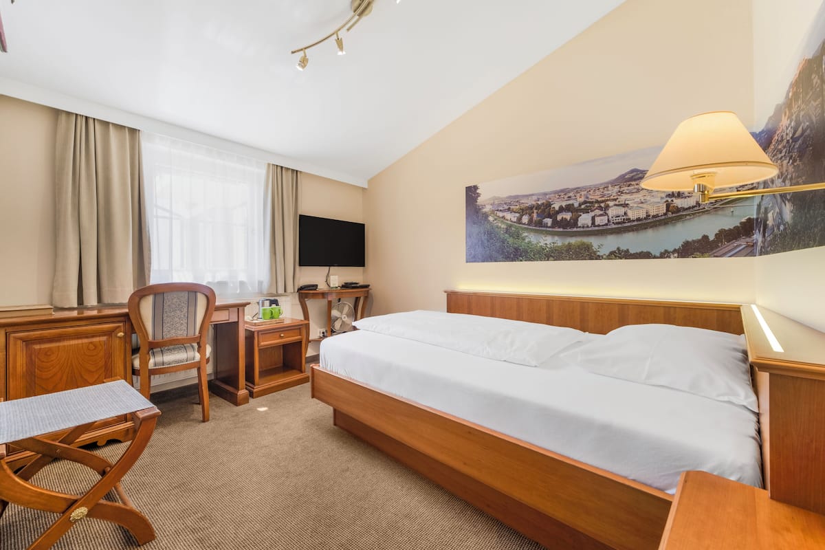 Quarto de hotel com cama, escrivaninha, cadeira, televisão e luminária fixada na parede.