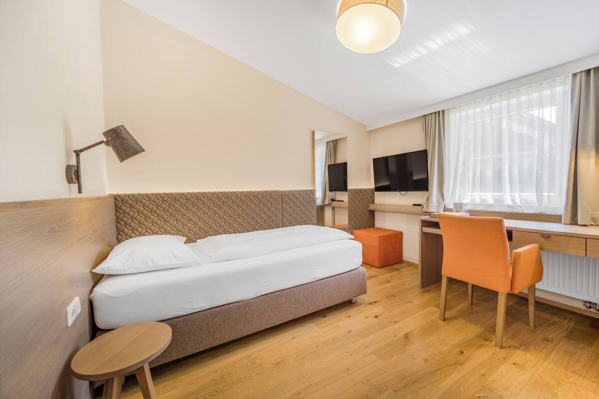 Quarto de hotel com cama, escrivaninha, cadeira, televisão e janela com cortinas.