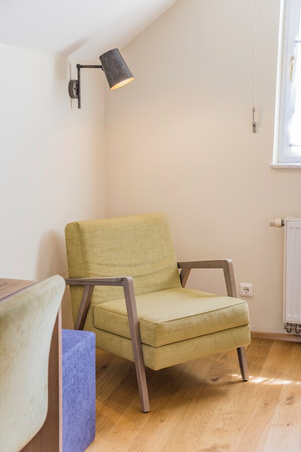 A room with a green upholstered chair, a wooden table, a wall-mounted lamp, and a window with blinds.