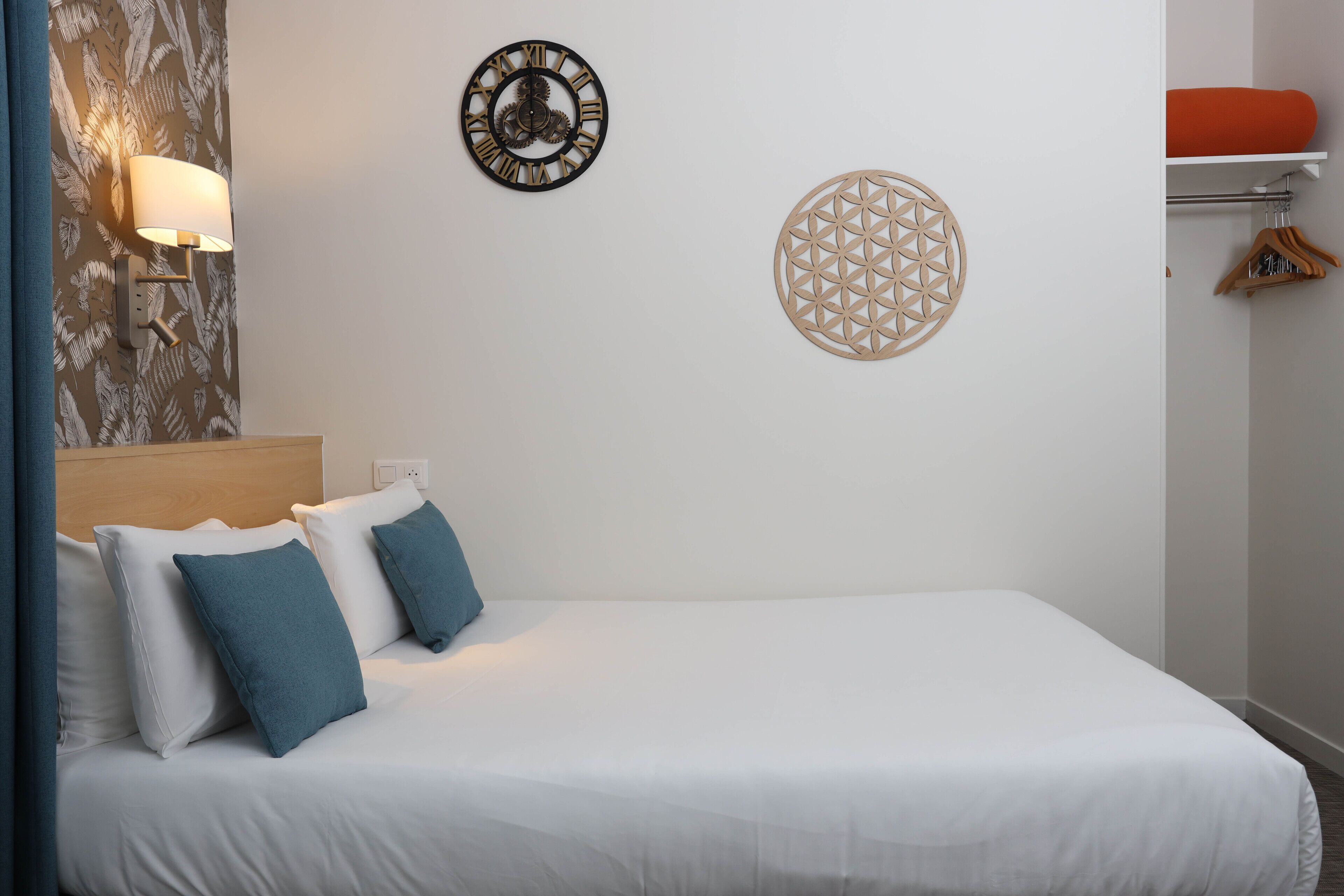 A neatly made bed with white and teal pillows, a wall clock, and a geometric wall decoration.
