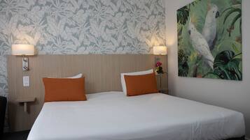 A hotel room with a large bed, two orange pillows, a floral wallpaper, and a painting of parrots.
