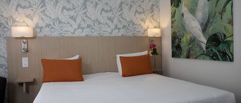 Una habitación de hotel con una cama grande, dos almohadas naranjas, un papel tapiz floral y un cuadro de loros.